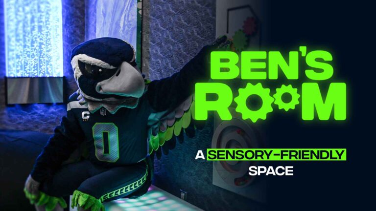 A costumed bird mascot sits in a dimly lit, sensory-friendly room with textured walls and soft lighting. The words "Ben's Room: A Sensory-Friendly Space" are prominently displayed in neon green text.