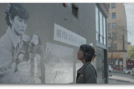 A person stands on a city street, looking at a large mural on a wall. The mural features a black and white image of a woman holding a child, with the words "Never Again Is Now" next to it. Buildings are visible in the background.