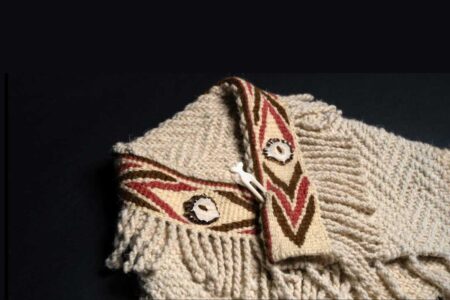 A close-up of a woven cream-colored garment with fringe, adorned with patterned bands in red, tan, and brown, featuring circular and chevron designs, and fastened with a small white bone toggle.