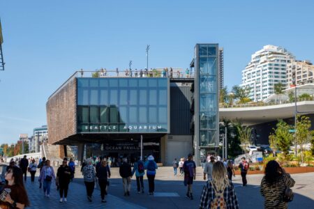 Crowds of people walk in front of the Seattle Aquarium’s Ocean Pavilion on a sunny day. The modern building has large glass windows, and people are visible on an upper lookout level. Tall buildings are in the background.