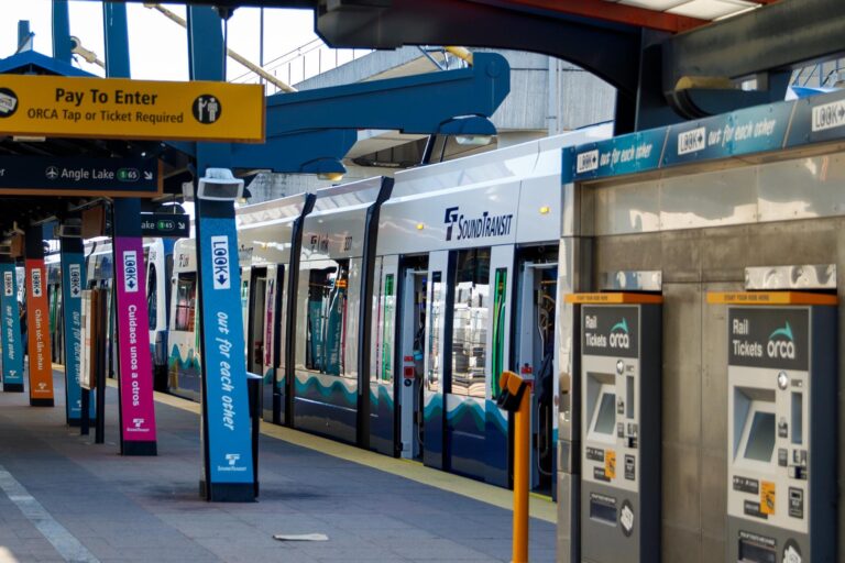 A Sound Transit light rail train at a station platform, with ticket machines on the right, colorful signs with rider information on pillars, and a yellow sign overhead indicating where to pay and tap an ORCA card.