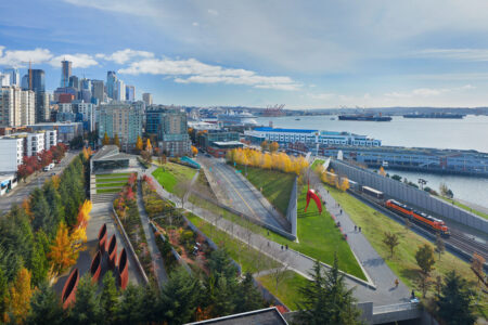 Aerial view of a cityscape featuring a waterfront park with green spaces, winding paths, and sculptures. Skyscrapers rise in the background, and a train passes by on tracks next to the water. The sky is partly cloudy with sunlight illuminating the scene.