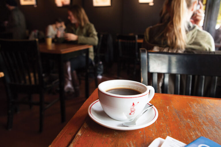 A cup of coffee sits on a wooden table in a cozy cafe. The background shows people sitting and chatting at tables, with soft lighting creating a warm atmosphere. A spoon rests on the saucer beside the cup.
