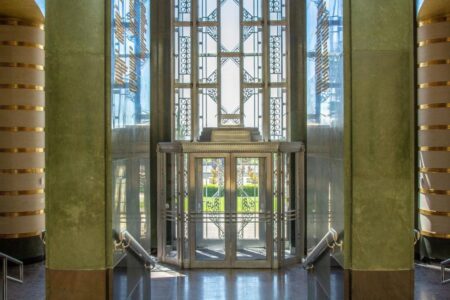 Art deco-style indoor entrance with ornate glass doors and intricate metalwork design. The walls and columns are green with gold accents, and a glimpse of sunny outdoors is visible through the glass, framed by geometric patterns.