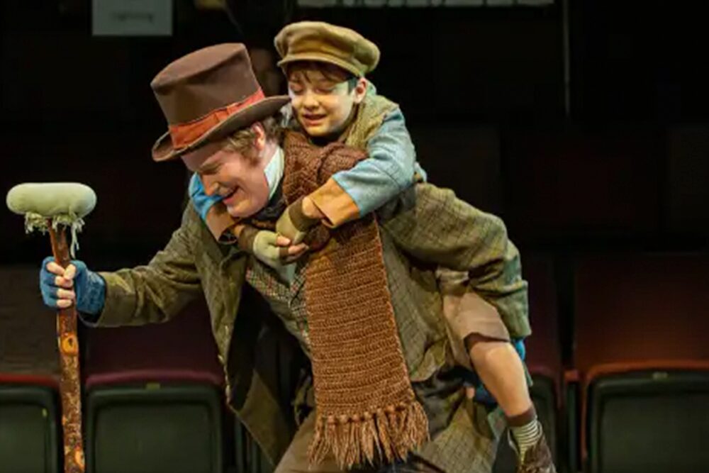A person wearing a brown top hat and scarf carries a child on their back. The child is dressed in vintage-style clothing, including a flat cap. Both appear joyful, and the setting looks like a theater stage.