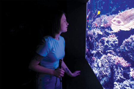 A person with a cane stands in low light, looking at a vibrant aquarium display filled with coral and colorful fish.