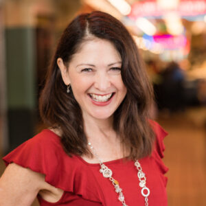 A woman with long brown hair and a big smile is wearing a red blouse and a silver necklace. The background is softly blurred with colorful lights, suggesting an indoor setting.