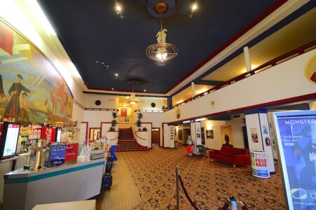 The interior of an ornate theater lobby features vintage decor, a concession stand with snacks and drinks, a staircase leading to an upper level, and murals on the walls. Plush carpeting and red seating add to the classic ambiance.