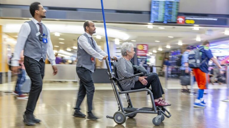 An airport employee pushes an elderly woman in a wheelchair through a busy terminal. Another employee walks alongside, both wearing uniforms. The background shows digital flight information screens and travelers.