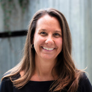 A woman with long brown hair smiles at the camera. She is wearing a black top and hoop earrings. The background features a blurred, muted interior with greenery.