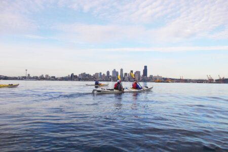 Two kayakers paddle on a calm body of water with the West Seattle neighborhood skyline in the background. The sky is partly cloudy, and other kayakers can be seen in the distance, creating a scene that conveys a peaceful morning on the water.