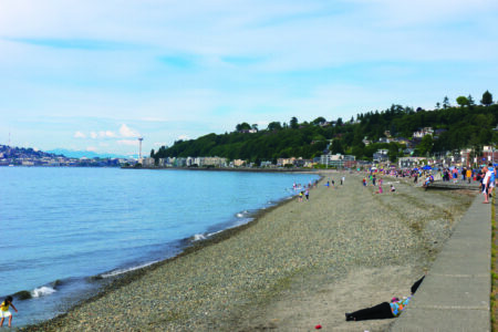 A scenic view of a pebble beach with people walking and enjoying the seaside. The water is calm, and a green hillside with houses is visible in the background under a partly cloudy sky.