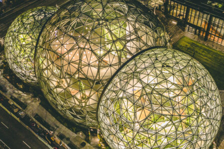 Aerial view of large, interconnected glass spheres with geometric patterns, illuminated from within. They house lush greenery, set in an urban environment with streets and buildings surrounding them at night.