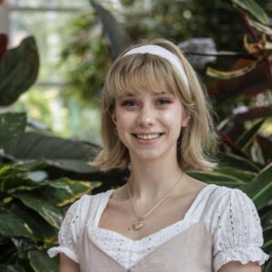 A young person with short blonde hair, wearing a white headband and a light pink dress over a white blouse, smiles in an indoor garden setting with lush green plants in the background.