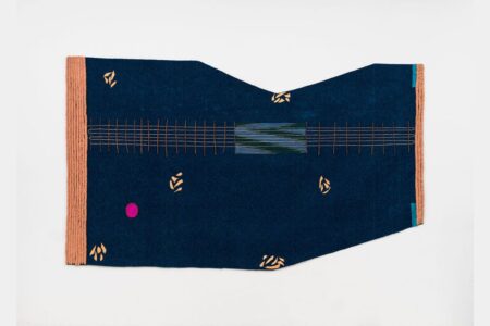 A blue, irregularly-shaped textile with small yellow patterns, a pink dot, a central rectangular design with horizontal stripes, tan ribbed edging on the left, and two small turquoise accents on the right edge.