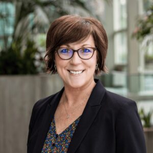 Woman with short brown hair and glasses, wearing a black blazer over a floral patterned blouse, smiles at the camera. The background shows indoor greenery and blurred architectural features.