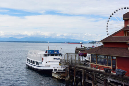 A scenic view of a waterfront with a sightseeing boat docked beside a pier. A large ferris wheel is visible in the background, and mountains can be seen across the water under a partly cloudy sky.
