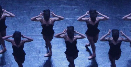 A group of dancers in black outfits perform in synchronized rows on a dimly lit stage. They stand with their feet apart, holding their arms bent upwards with hands near their heads, creating a symmetrical pattern.
