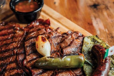 Grilled steak arranged on a wooden board with roasted onion, jalapeño, and sausage captures the essence of a hearty meal. A small black cup of sauce waits in the background as if at home on a rustic table in the Ballard neighborhood.