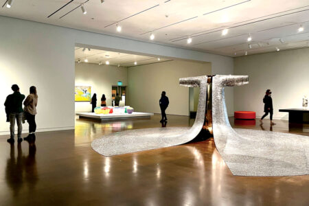 A spacious art gallery with polished floors features a large metallic sculpture in the center. People are observing various artworks on the walls and a colorful installation on a platform. The room is well-lit with overhead lights.