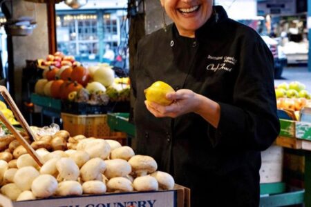 A smiling woman in a black chef's coat stands by a vibrant market stall. She holds a lemon and is surrounded by fresh produce, including tomatoes and mushrooms. The setting is lively, with people and market signs in the background.