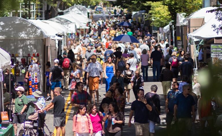 A crowded street fair with numerous people walking along tents and stalls. Some are shopping, others chatting, and a few holding umbrellas. Trees line the street, providing shade on a sunny day.
