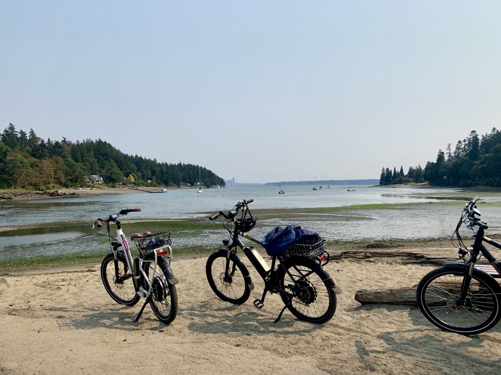 Three bicycles are parked on a sandy beach with a calm bay and several small boats in the water. Trees line the shore on both sides under a clear sky.