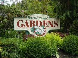 Sign reading "Bainbridge Gardens" surrounded by lush greenery and shrubs. A large flower illustration decorates the center of the sign.