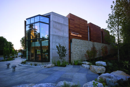 The modern art museum, nestled between Bainbridge Island and Bremerton, features large glass windows and a striking concrete and wood facade. It is beautifully surrounded by landscaped greenery and a charming stone pathway.