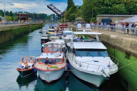 A busy waterway in the Ballard neighborhood features various boats aligned closely, navigating through a canal. A raised drawbridge and lush greenery are visible in the background. People gather along the canal banks, observing the scene under a partly cloudy sky.