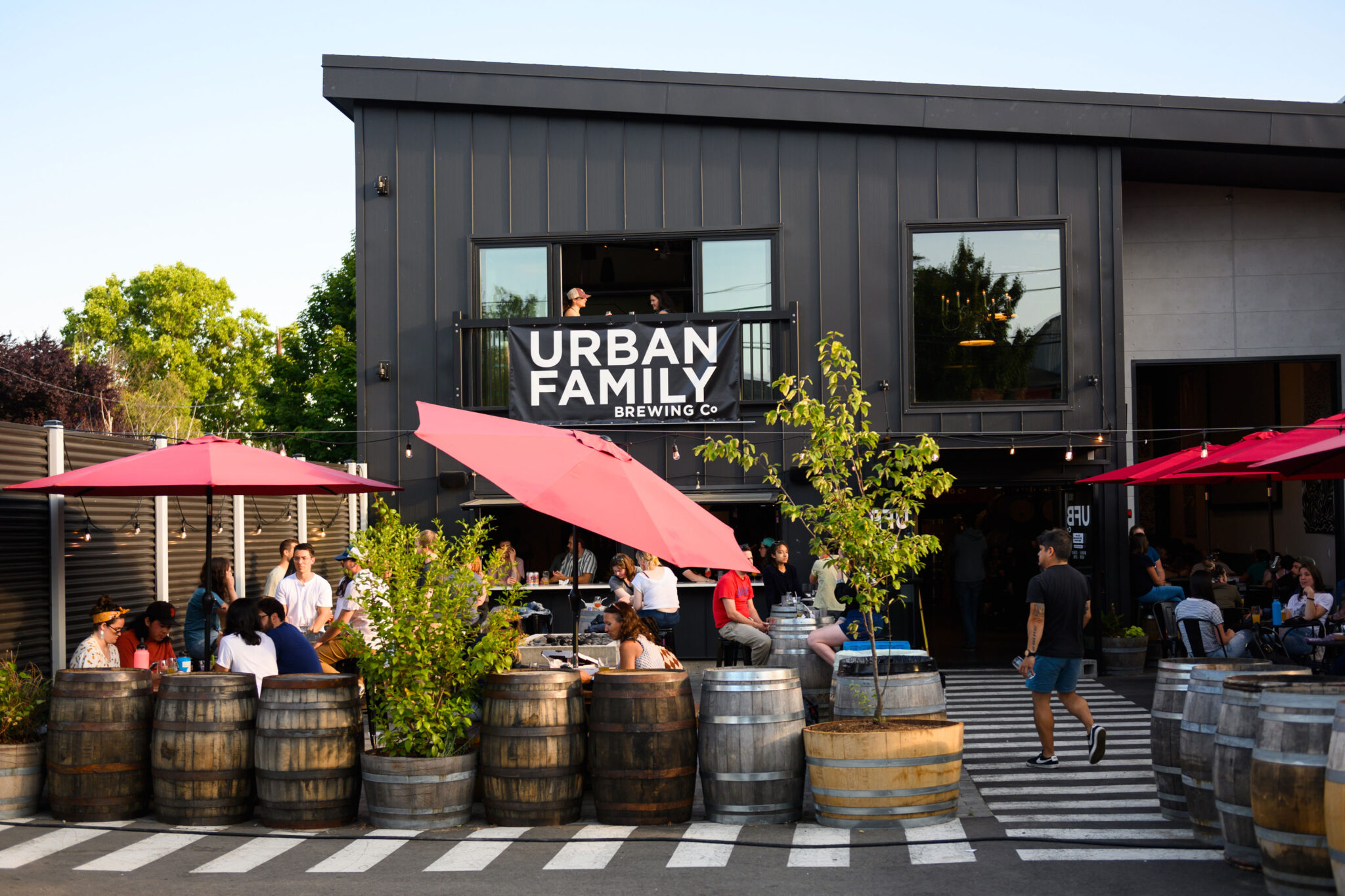 A lively brewery setting with people sitting under pink umbrellas surrounded by barrels. The black building has a sign reading "Urban Family Brewing Co." Trees and plants add greenery to the scene.