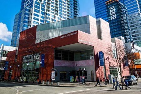 Street view of the Bellevue Arts Museum, showcasing a modern red brick facade and large windows. Tall buildings surround the museum, and a few people walk along the sidewalk. Trees and banners can be seen in the foreground.