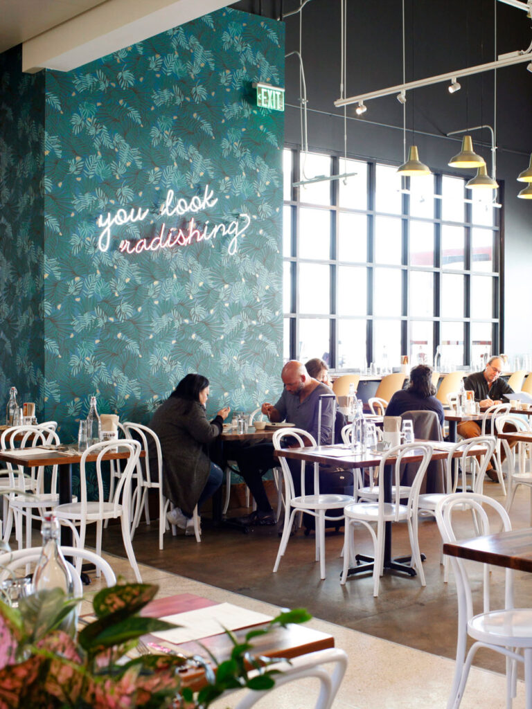 People sitting at white tables and chairs fill a room with a blue wall and windows in the background.