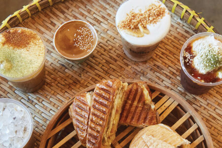 Five coffee beverages and five sandwiches sit on a woven tabletop.