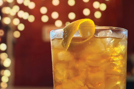 A glass filled with an iced amber-colored drink garnished with a lemon twist. The background features warm, blurred circular lights, creating a cozy atmosphere.