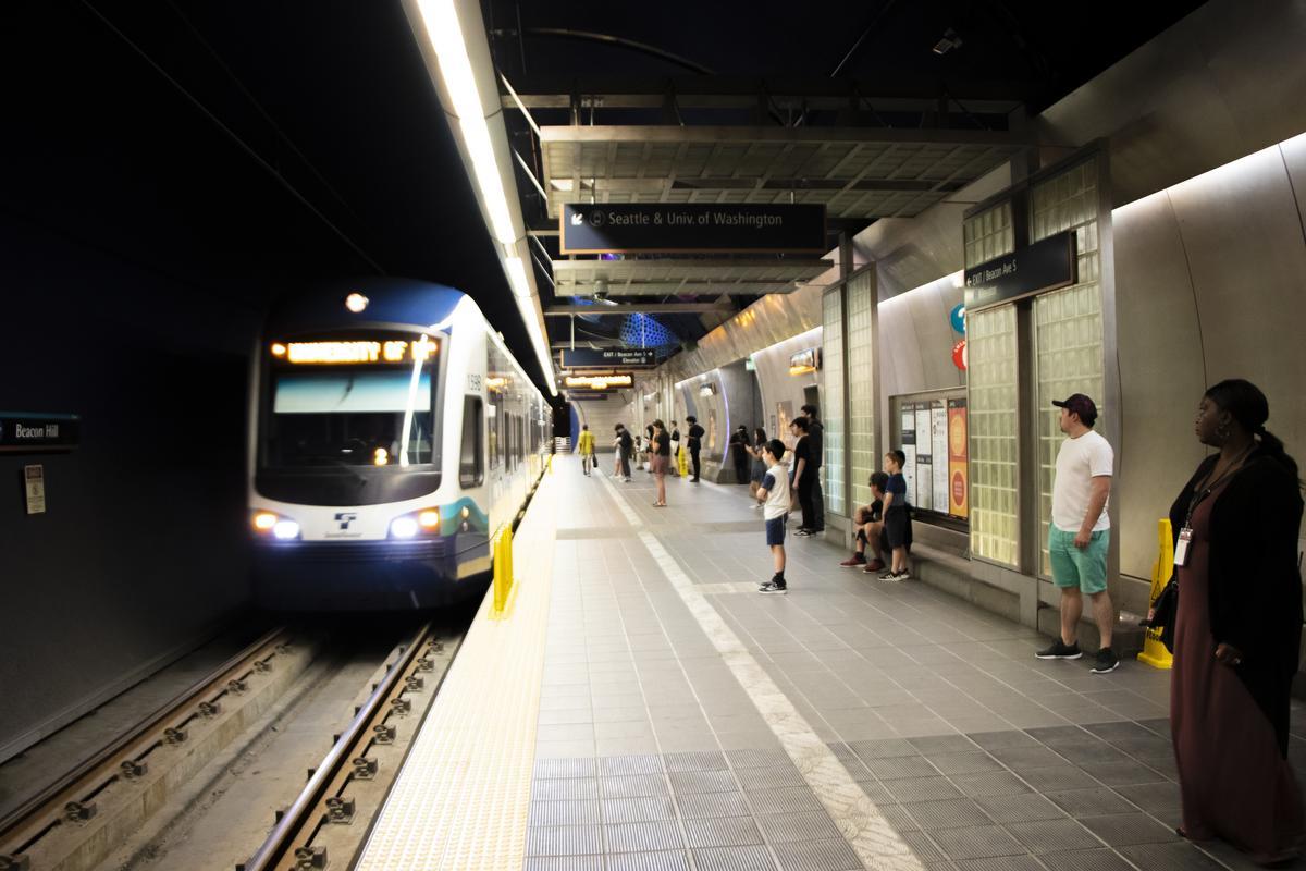 A blue and white light rail train arrives at an underground station as people wait on the platform. Some passengers sit on benches while others stand near the edge, ready to board. Signs indicate directions to Seattle and the University of Washington.