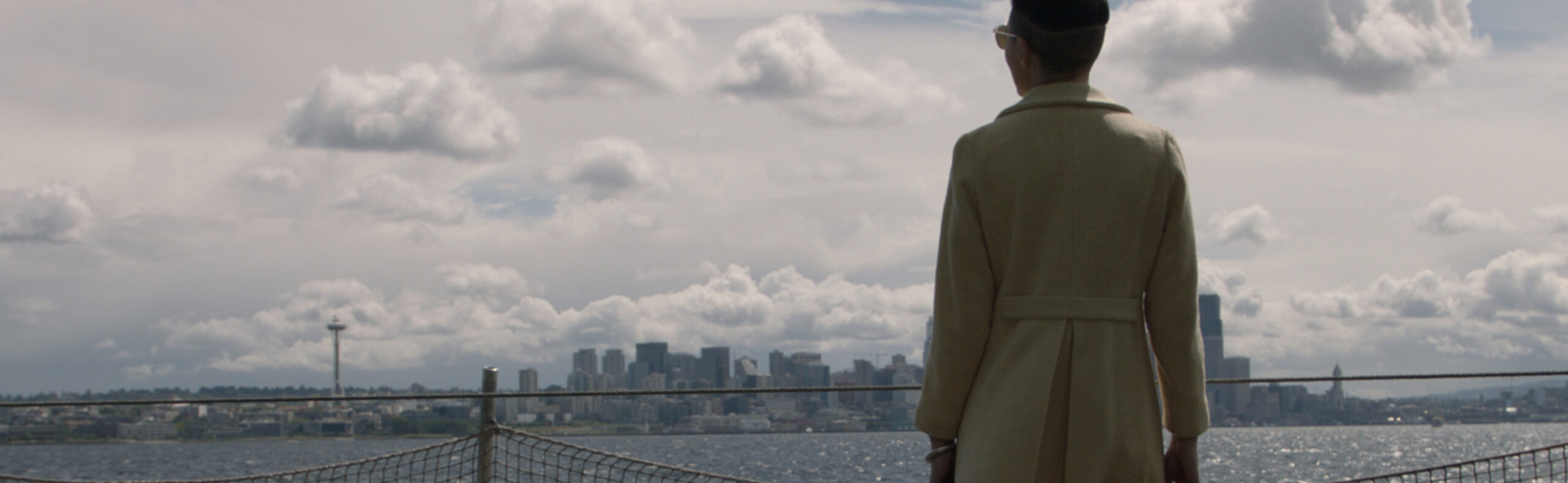 A person in a trench coat and hat stands on a boat, facing a city skyline across a body of water. The sky is overcast with scattered clouds, and a distinctive tower is visible on the left side of the skyline.
