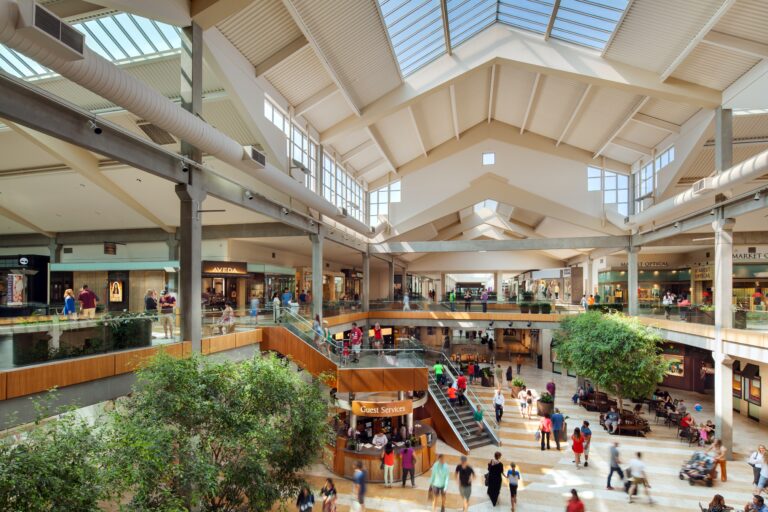 A busy, modern shopping mall with multiple floors and a skylight ceiling. Shoppers walk and gather around a coffee stand, while others explore stores on both levels. Green plants decorate the open space, adding a touch of nature.