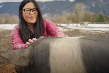 A person with long dark hair and glasses is smiling while petting a pig. They are outdoors in a snowy, mountainous area. The pig has a black and white coat.