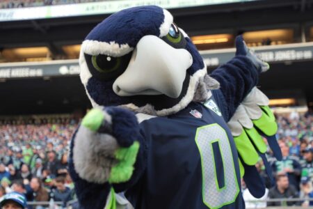 A large, blue and green bird mascot is wearing a sports jersey with the number 0, posing energetically in an open stadium filled with fans. The mascot's wings are outstretched, and it appears to be engaging with the crowd.