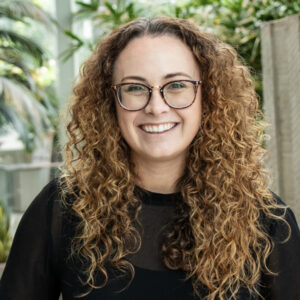 A person with long curly hair and glasses smiles at the camera. They are wearing a black top. The background features lush green plants and natural light.
