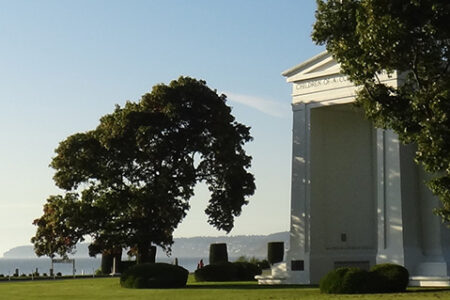 A white monument stands beside a large tree in a grassy area. The sky is clear and blue, and the ocean can be seen in the background. Bushes and distant land are visible beyond the water.