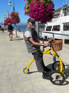 A person using a yellow mobility aid on a sidewalk lined with pink hanging flowers with a white boat on the righthand side.