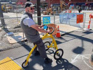 A person using a yellow mobility aid sits in the foreground in front of a chainlink fence and construction work in the background.