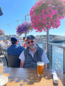 A person in a grey vest sitting at a wood table with a beer on top. Pink and purple flowers hang overhead and water is seen on the righthand side.