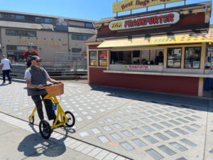 A person using a yellow mobility aid sits on the sidewalk in front of a red building with yellow awning.