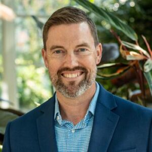 Smiling man with short hair and a beard wearing a blue suit and checkered shirt, standing outdoors with greenery in the background.