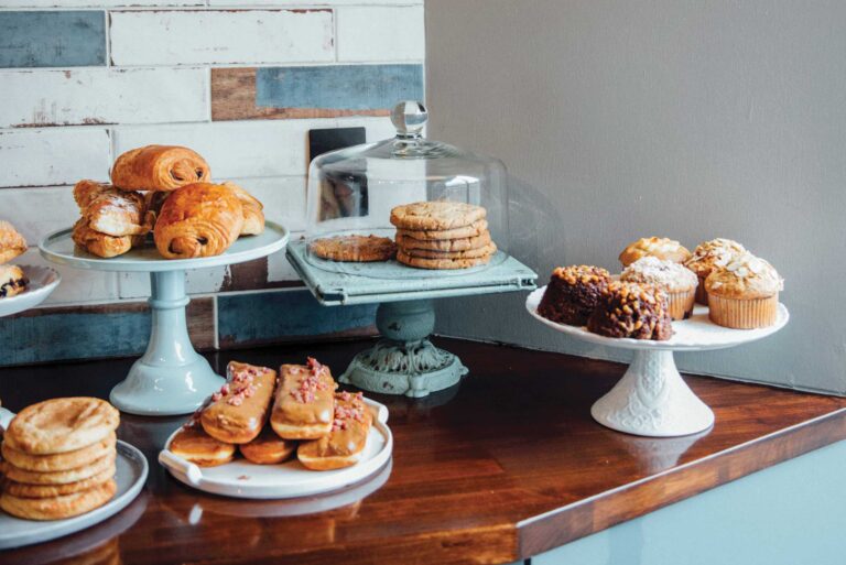 A display of assorted pastries on a wooden counter, including croissants, muffins, cookies, and eclairs. Some are placed on decorative stands, while others are arranged on plates or under a glass dome. A white and brick-patterned wall is in the background.