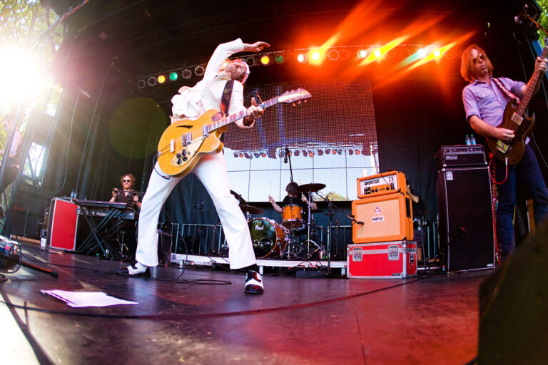 A musician in a white suit energetically plays an electric guitar on stage, with a band in the background, as one of the many exciting things to do in Seattle. Bright stage lights and sunlight create a vibrant atmosphere, with a keyboard player, drummer, and bassist enhancing the lively performance.