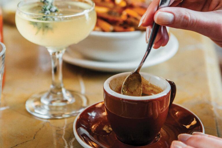 A person stirs a small cup of espresso on a brown saucer, next to a cocktail garnished with herbs. A bowl of food is blurred in the background, all set on a marble table.